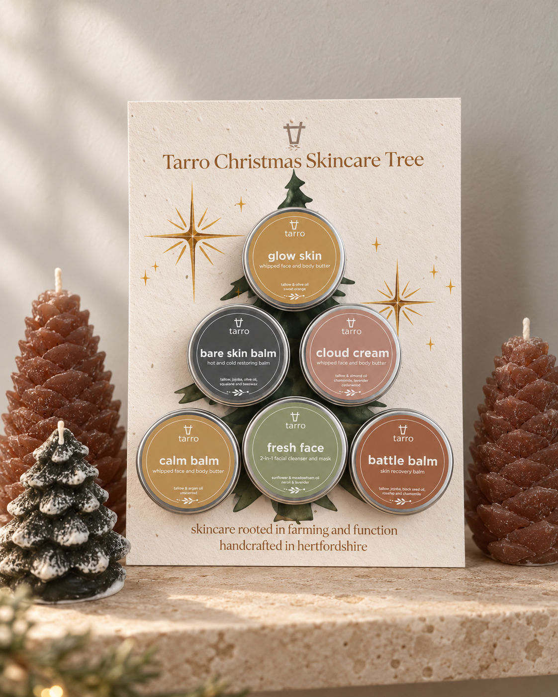 A set of six Tarrto skincare balm tins arranged in a Christmas tree shape on a branded display card, styled with decorative pinecones and a small snow-dusted tree on a stone surface.