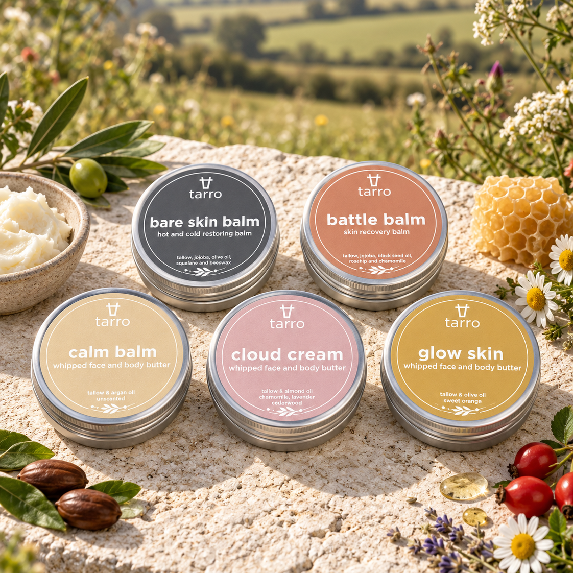 Five round metal tins of tarro body balm and cream are arranged on a stone surface outdoors, surrounded by flowers, leaves, olives, honeycomb, and small berries. The tins have different colored labels: gray, peach, beige, pink, and gold, with labels reading bare skin balm, battle balm, calm balm, cloud cream, and glow skin.